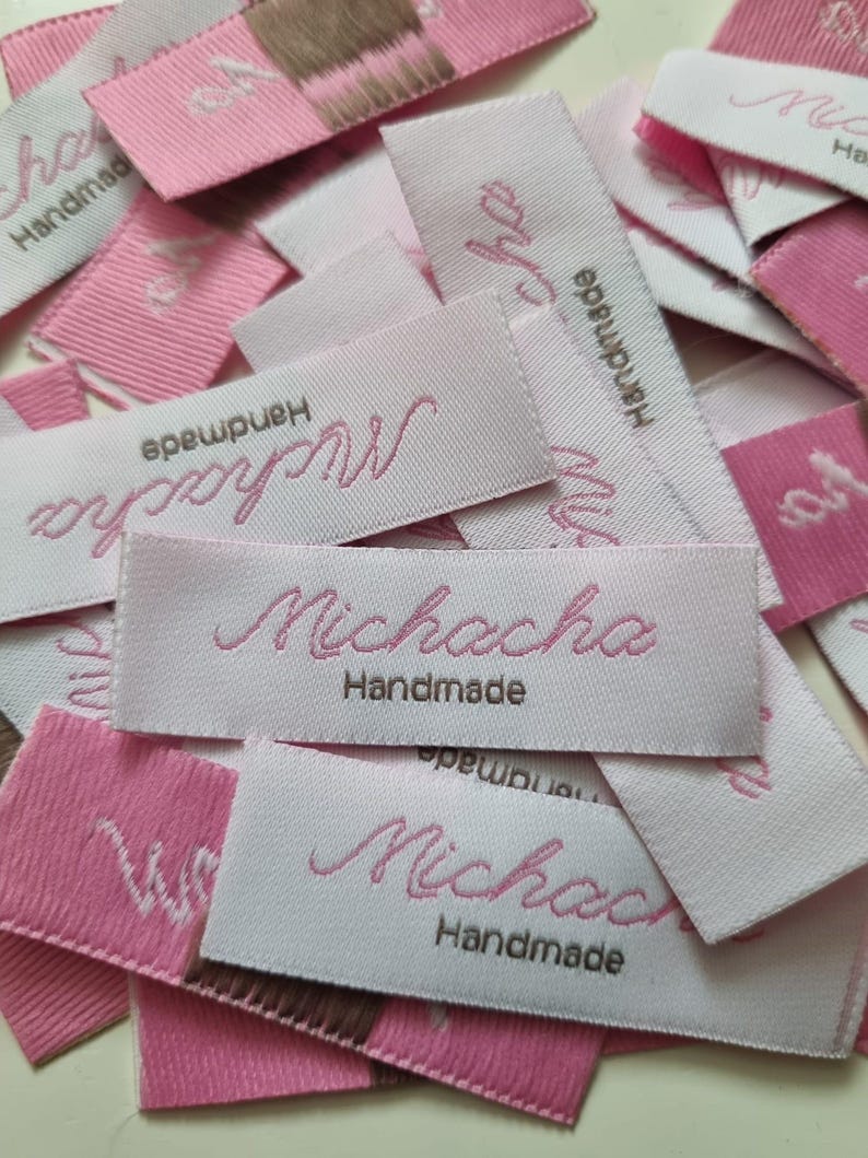 Pu&ograve; includere: Una collezione di etichette rettangolari in tessuto per abbigliamento nei toniamento nei toni del rosa chiaro e del bianco. Ogni etichetta presenta la parola Michacha ricamata in corsivo rosa, con la parola Handmade cucita in filo marrone sotto.