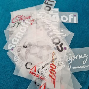 May include: A stack of clear plastic sheets with various red, black, and white text printed on them. The text includes "Sanofi", "Golden 007", "Sensations", "Proje Okul", "C", "A", "R", "Tyoruz", and "Enerjisaoret".