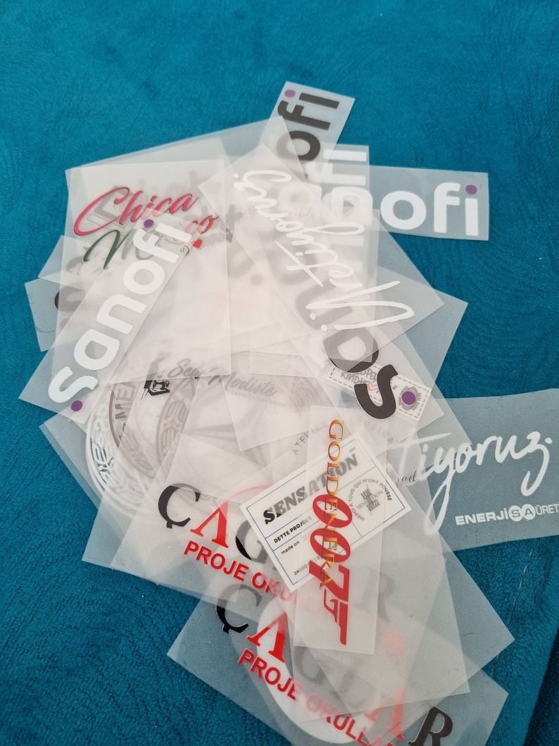 May include: A stack of clear plastic sheets with various red, black, and white text printed on them. The text includes "Sanofi", "Golden 007", "Sensations", "Proje Okul", "C", "A", "R", "Tyoruz", and "Enerjisaoret".