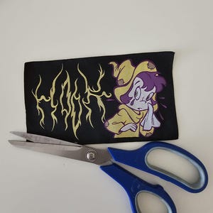 Pu&ograve; includere: Una toppa rettangolare in tessuto nero con una grafica di testo gialla stilizzata che recita "HOOK" e un'illustrazione a cartone animato di un personaggio dai capelli viola che indossa un mantello giallo con cappuccio, sopra un paio di forbici.