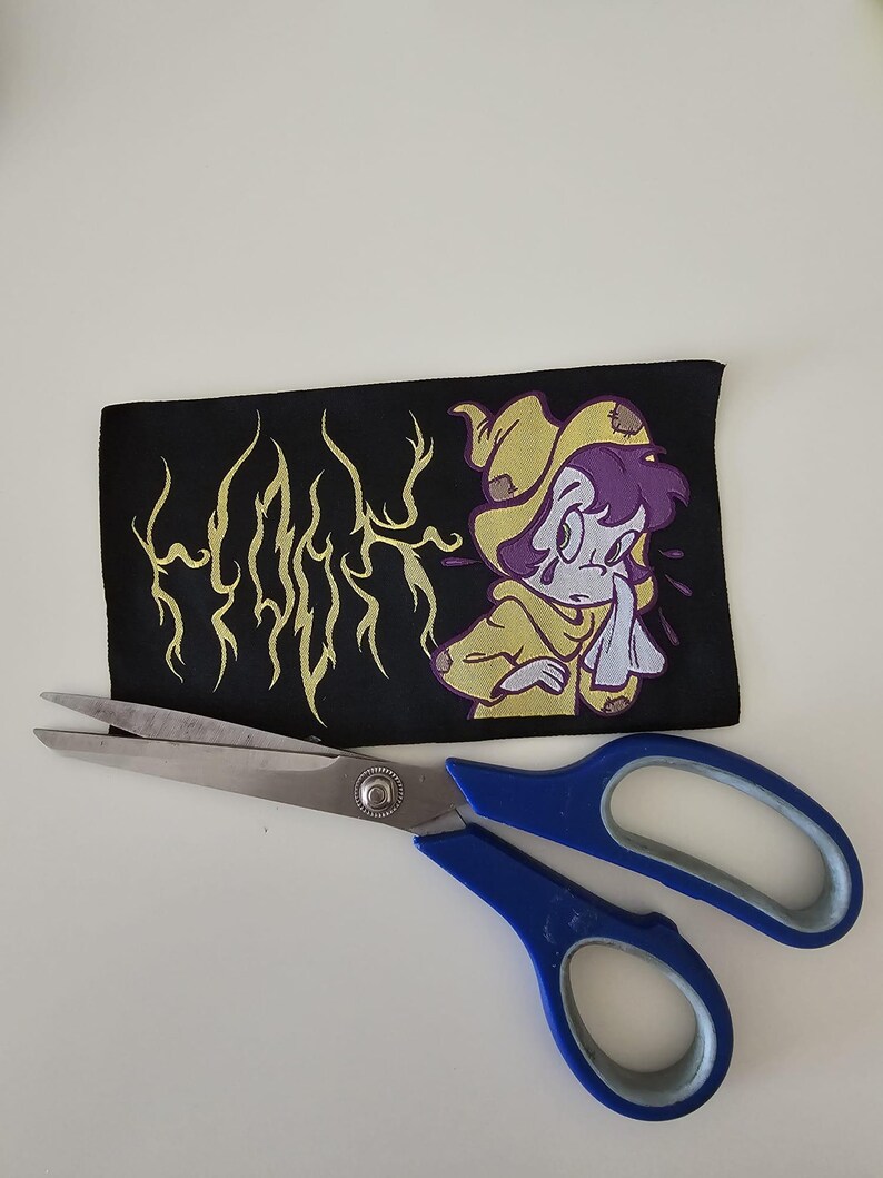 Pu&ograve; includere: Una toppa rettangolare in tessuto nero con una grafica di testo gialla stilizzata che recita "HOOK" e un'illustrazione a cartone animato di un personaggio dai capelli viola che indossa un mantello giallo con cappuccio, sopra un paio di forbici.