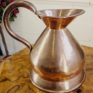 May include: A polished copper pitcher featuring a wide, rounded body, a flared rim, and a curved handle. The metal surface exhibits a warm, metallic sheen with subtle reflections, showcasing a smooth, seamless construction from the base to the top.