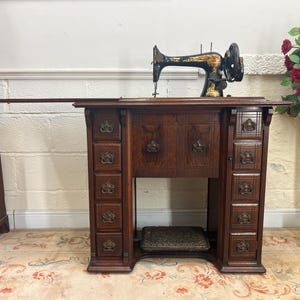 May include: A vintage treadle sewing machine housed in a dark wood cabinet with ten small drawers featuring ornate metal pulls. The black sewing machine head has gold decorative accents and sits atop the cabinet, which includes a foot pedal.