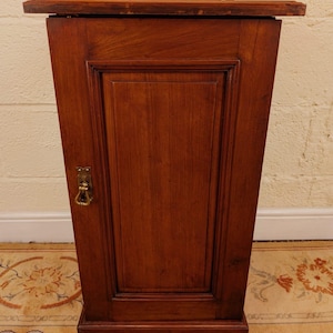 May include: An antique wooden cabinet in a deep reddish-brown hue. The cabinet has a single door with a recessed panel and a brass handle. The top is adorned with carved detailing. It stands on a patterned rug, showcasing its vintage design. The cabinet's height is approximately 100 cm.