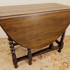 May include: A dark brown wooden drop-leaf table featuring a rectangular top with rounded edges and two hinged side leaves. The table is supported by four barley twist legs connected by a rectangular base stretcher frame with block feet.