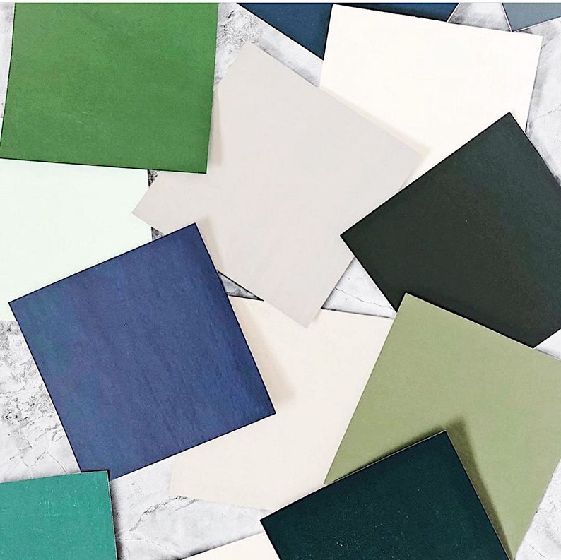 May include: An assortment of square-shaped paper samples in various shades of green, blue, and neutral tones. The colors range from deep forest green to pale sage, with a mix of navy, light gray, and cream. The squares are scattered on a light-colored surface.