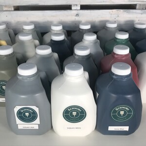 May include: A collection of paint jugs in various colors, including gray, white, and navy blue. Each jug has a white lid and a label with the brand name "Schnurs". The paint colors are labeled "Volcanic Ash", "Delicate White", and "Navy Blue".