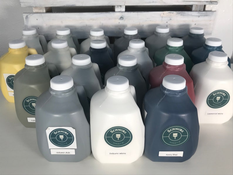 May include: A collection of paint jugs in various colors, including gray, white, and navy blue. Each jug has a white lid and a label with the brand name "Schnurs". The paint colors are labeled "Volcanic Ash", "Delicate White", and "Navy Blue".