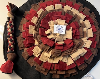 May include: A dog puzzle mat with a black base and a circular design of red, brown, and tan felt squares. A braided rope toy with red and brown accents is attached to the side. A small white tag is visible.