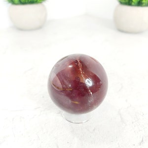 Puede incluir: Una esfera pulida de color rojo y dorado, probablemente de ágata, se encuentra sobre un soporte transparente. La esfera tiene un acabado liso y brillante y patrones intrincados de rojo y dorado.