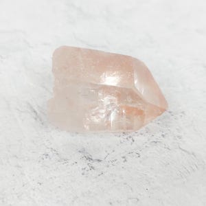 Könnte beinhalten: Ein klarer, rosa Quarzkristall mit einer spitzen Spitze. Der Kristall hat eine natürliche, raue Textur und ist durchscheinend.