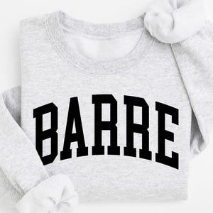 May include: A light gray sweatshirt with the word "BARRE" printed in bold, black, block letters across the front. The sleeves are folded, and the overall look is casual and comfortable. Perfect for fitness enthusiasts.