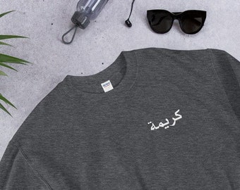 May include: A dark gray sweatshirt with the Arabic word "Kareema" embroidered in white on the chest. The sweatshirt is laid flat on a textured surface, with sunglasses, a water bottle, and a phone nearby.