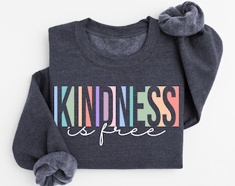 Könnte beinhalten: Ein dunkelgraues Sweatshirt mit dem Aufdruck "KINDNESS is free" in bunten Blockbuchstaben und Schreibschrift. Die langen Ärmel sind umgeschlagen, und das Sweatshirt ist gefaltet.
