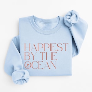 May include: Light blue sweatshirt with the text "Happiest by the Ocean" in coral pink lettering. The sweatshirt has a palm tree graphic.