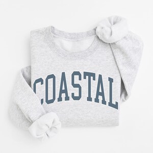 May include: A light gray sweatshirt with the word "COASTAL" printed in a dark blue, arched font across the chest. The sleeves are folded, and the collar is visible. The sweatshirt is made of a soft material.