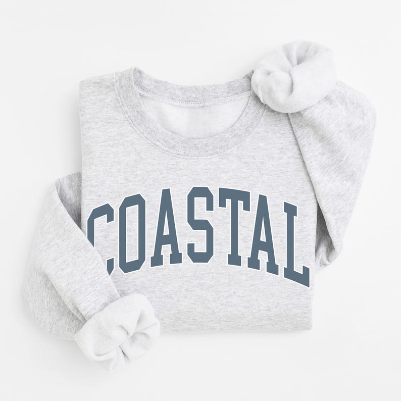 May include: A light gray sweatshirt with the word "COASTAL" printed in a dark blue, arched font across the chest. The sleeves are folded, and the collar is visible. The sweatshirt is made of a soft material.