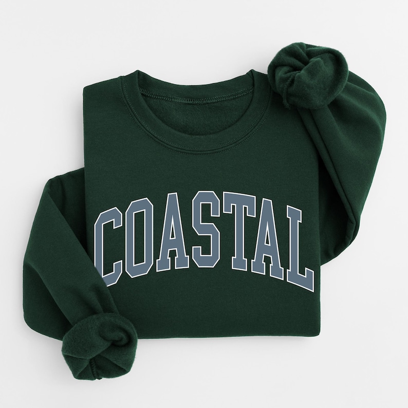 May include: A dark green sweatshirt with the word "COASTAL" in large, block letters across the front. The letters are white with a gray outline. The sweatshirt is folded, with one sleeve bunched up.