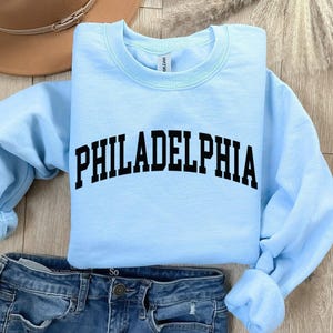 May include: Light blue sweatshirt with the text "PHILADELPHIA" in black, arched lettering. The sweatshirt is shown on a pair of blue jeans.
