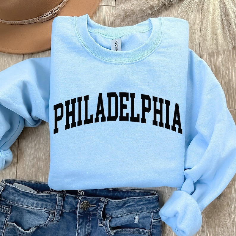 May include: Light blue sweatshirt with the text "PHILADELPHIA" in black, arched lettering. The sweatshirt is shown on a pair of blue jeans.