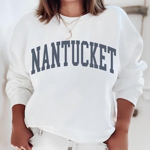 Könnte beinhalten: Ein weißes Sweatshirt mit Rundhalsausschnitt und dem Wort NANTUCKET, das in einer großen, geschwungenen, dunkelgrauen Serifenschrift auf der Brust gedruckt ist. Das Kleidungsstück hat lange Ärmel und einen gerippten Ausschnitt.