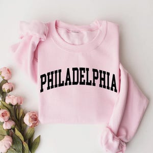 May include: A pink sweatshirt with the text "PHILADELPHIA" printed in black across the front.