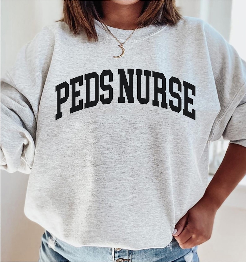 Peut inclure: Un sweat-shirt gris avec le texte "PEDS NURSE" en lettres noires en arc de cercle.