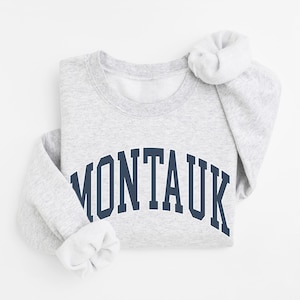 Puede incluir: Una sudadera de cuello redondo en color gris jaspeado claro con la palabra MONTAUK impresa en azul marino oscuro con una tipografía de estilo universitario en el pecho. La prenda tiene mangas largas con puños acanalados y un cuello redondo acanalado.