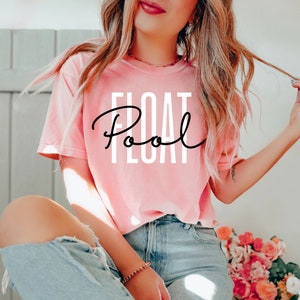 Könnte beinhalten: Ein pinkfarbenes T-Shirt mit dem Text "Float Pool" in schwarzer Schrift auf der Vorderseite. Das Shirt wird von einer Person mit langen blonden Haaren getragen und zu einer blauen Jeans kombiniert.