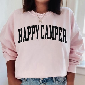 Peut inclure: Un sweat-shirt rose avec le texte "HAPPY CAMPER" en lettres noires.