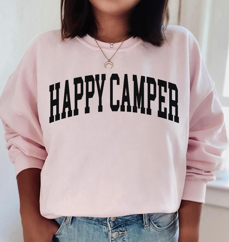 Peut inclure: Un sweat-shirt rose avec le texte "HAPPY CAMPER" en lettres noires.
