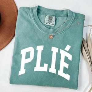 May include: A teal Comfort Colors t-shirt with the word "PLIÉ" in large white block letters. A silver necklace with a small pendant rests on the shirt. The shirt is folded and placed on a white surface.