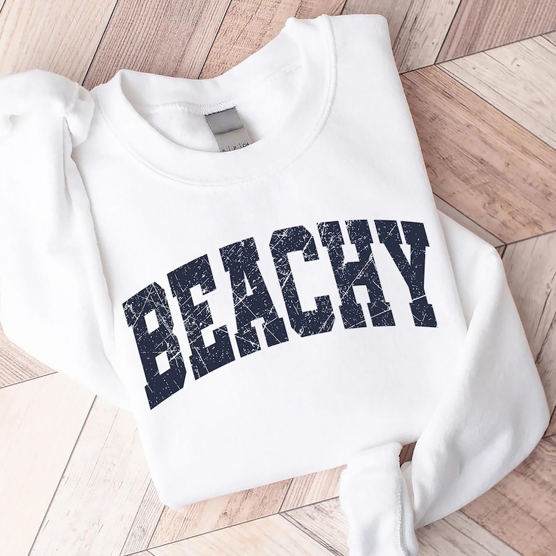 May include: White sweatshirt with a navy blue distressed graphic that says "Beachy" in a collegiate font.