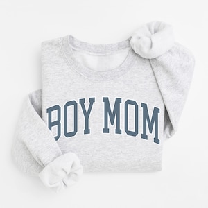 May include: A light gray sweatshirt with the text "BOY MOM" in a blue collegiate font. The sleeves are folded, and the sweatshirt is neatly arranged. The overall aesthetic is casual and comfortable.