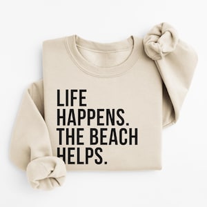 May include: A beige sweatshirt with the text "LIFE HAPPENS. THE BEACH HELPS." printed in black.
