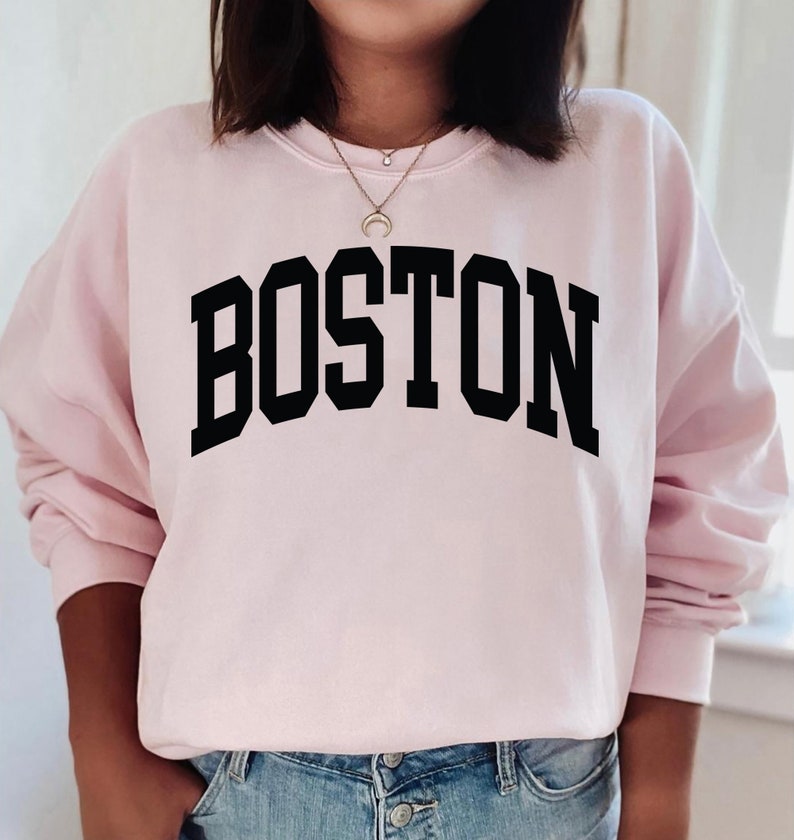 K&ouml;nnte beinhalten: Ein rosa Sweatshirt mit dem Wort "BOSTON" in schwarzen Blockbuchstaben.