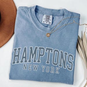 May include: A blue short-sleeved t-shirt with a crew neckline and a Comfort Colors brand tag. The shirt features the white text HAMPTONS NEW YORK printed in a collegiate font across the chest. A gold-toned necklace with a small circular pendant rests on the fabric.