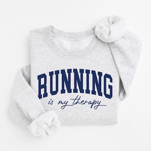 Puede incluir: Sudadera gris claro con el texto "RUNNING is my therapy" en azul marino. La palabra "RUNNING" está en negrita, en forma de arco, mientras que "is my therapy" está en cursiva. Las mangas están dobladas.
