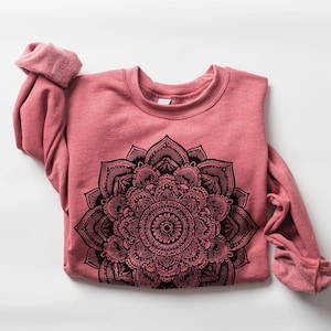 May include: A dusty rose-colored sweatshirt with a crew neck and long sleeves. The front of the sweatshirt features a large, intricate black mandala design. The sleeves are cuffed at the wrists, and the garment is folded.
