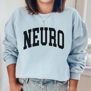 K&ouml;nnte beinhalten: Ein hellblauer Pullover mit dem Wort "NEURO" in schwarzer Schrift, in einer College-Schriftart.
