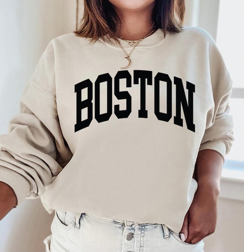 K&ouml;nnte beinhalten: Ein beiger Pullover mit dem Wort "BOSTON" in schwarzen Blockbuchstaben.
