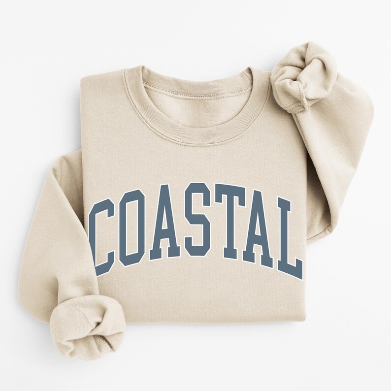 May include: A beige sweatshirt with the word "COASTAL" printed across the front in a blue, collegiate-style font. The sleeves are folded, and the sweatshirt is neatly arranged. The overall aesthetic is casual and comfortable.