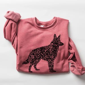 Peut inclure: Un sweat-shirt rose poudré avec un motif floral noir représentant un berger allemand. La silhouette du chien est remplie de motifs de fleurs et de feuilles. Le sweat-shirt a un col rond et des manches longues.