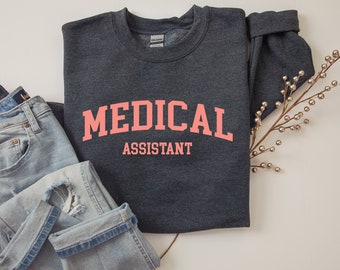 May include: A dark grey sweatshirt with the text "MEDICAL ASSISTANT" in pink lettering. The sweatshirt is folded and laid on a white surface.