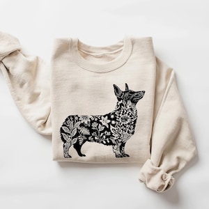 May include: Cream-colored sweatshirt featuring a black and white floral corgi design. The corgi's body is filled with detailed botanical illustrations. The sweatshirt has a crew neck and long sleeves, perfect for casual wear.