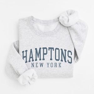 Puede incluir: Una sudadera de cuello redondo en color gris claro con el texto HAMPTONS NEW YORK impreso en una fuente azul oscuro de estilo universitario en el pecho. La prenda tiene mangas largas con puños acanalados y un cuello acanalado de diseño clásico.