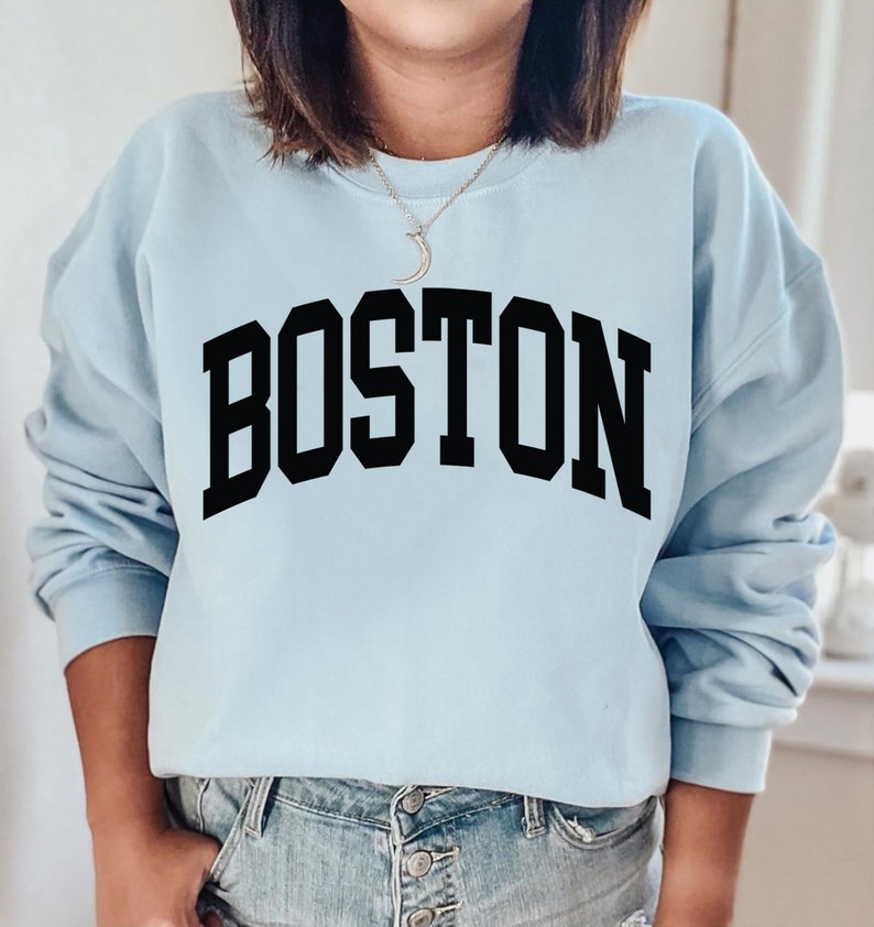 K&ouml;nnte beinhalten: Ein hellblauer Pullover mit dem Wort "BOSTON" in schwarzen Blockbuchstaben.
