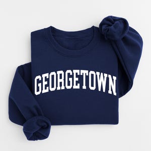 Peut inclure: Un sweat-shirt bleu marine avec le mot "GEORGETOWN" imprimé en blanc sur le devant. Les manches sont pliées. Le vêtement est en matière douce, idéal pour un usage quotidien.