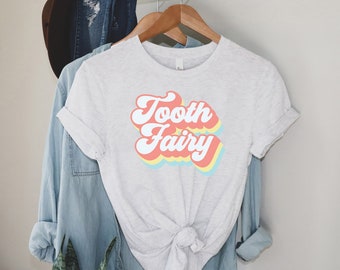 Może przedstawiać: Biała koszulka z grafiką w stylu retro, na której widnieje napis "Tooth Fairy" w kolorach tęczy.