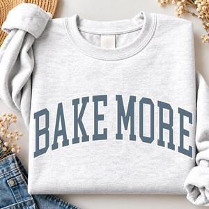 May include: A light gray crewneck sweatshirt featuring the text BAKE MORE in a dark blue collegiate-style font arched across the chest. The garment has long sleeves, a ribbed neckline, and a soft heathered texture throughout the fabric.